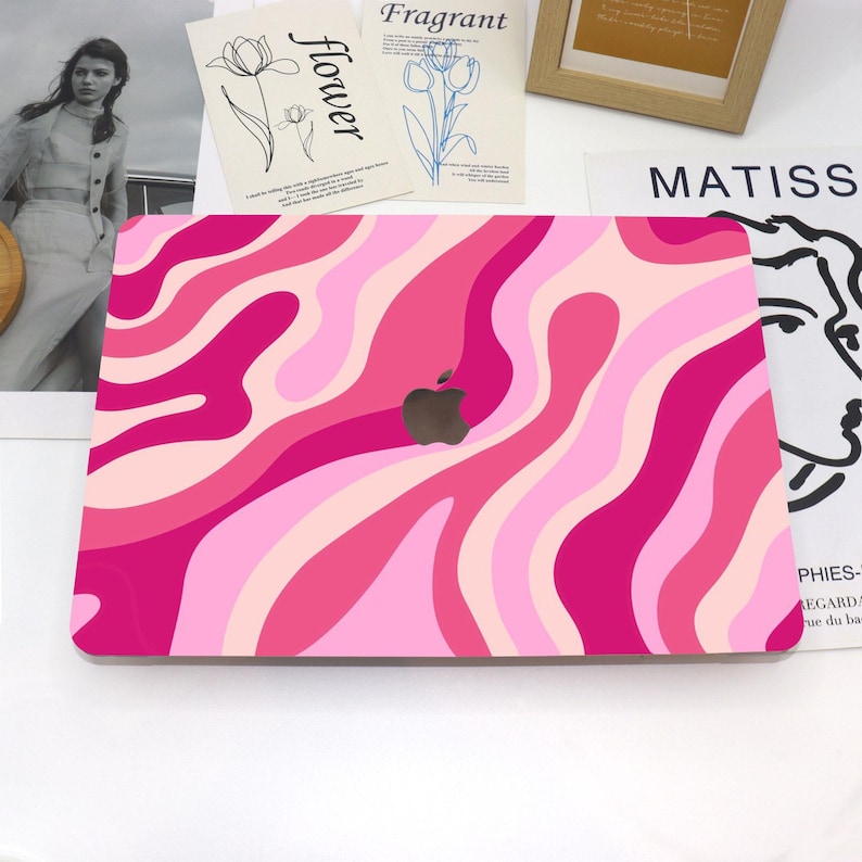 May include: A pink laptop case with a wavy pattern in shades of pink and light pink. The Apple logo is visible in the center. The case is on a white surface with decorative items in the background.