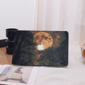 May include: Laptop case featuring a nighttime forest scene with a large, glowing moon and stars. The Apple logo is prominently displayed. The case has a dark background with silhouetted trees.