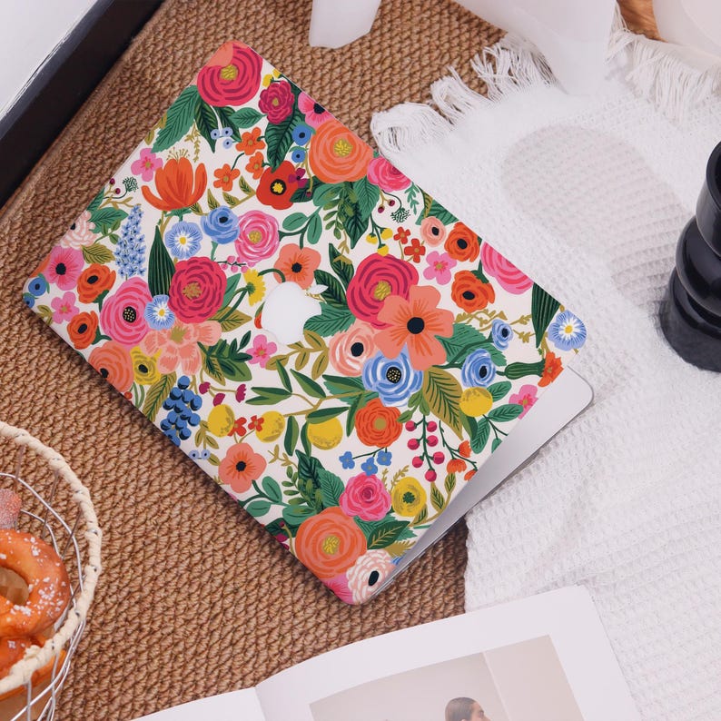 May include: A laptop case with a vibrant floral pattern featuring pink, orange, blue, and yellow flowers on a white background. The case has a cutout for the Apple logo. The design includes green leaves and stems, creating a colorful and cheerful aesthetic.