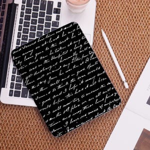 May include: A black tablet case featuring a white handwritten cursive script pattern across the entire surface. The case is rectangular with rounded corners and is positioned next to a white stylus on a laptop keyboard and a textured surface.