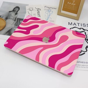 May include: A laptop case with a vibrant, wavy pattern in shades of pink. The case features a silver Apple logo and a smooth, glossy finish. The design includes various shades of pink, creating a retro, groovy aesthetic.