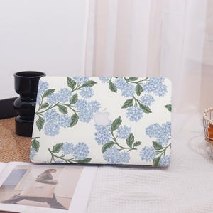 Pode incluir: Uma capa branca para laptop com um padrão floral de flores de hortênsia azul claro e folhas verdes. O logotipo da Apple é visível. A capa está em uma superfície branca.