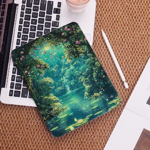 Peut inclure: Une coque de tablette avec une illustration vibrante d'une scène de forêt sereine avec une cascade, un lac et des fleurs roses. L'étui repose sur un ordinateur portable, avec un stylet blanc et une boisson à proximité. La palette de couleurs est verte et bleue.