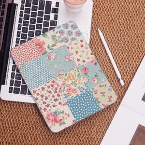 May include: A floral patchwork design tablet case featuring a variety of patterns and colors, including pink roses, blue polka dots, and floral prints. The case is resting on a laptop keyboard and a woven surface.