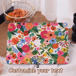 May include: A laptop case with a floral design featuring pink, orange, and blue flowers on a white background. The case has the Apple logo cutout and the name "Ashley" is written on the case. The text "Customize your text" is also visible.