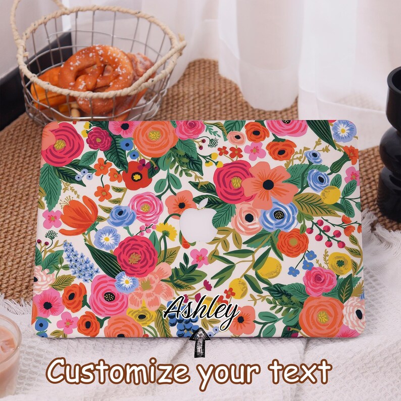 May include: A laptop case with a floral design featuring pink, orange, and blue flowers on a white background. The case has the Apple logo cutout and the name "Ashley" is written on the case. The text "Customize your text" is also visible.