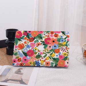 May include: A laptop case with a vibrant floral pattern. The design features a white background covered in colorful flowers, including pink, orange, and blue blooms, with green leaves. The case has a cutout for the Apple logo.