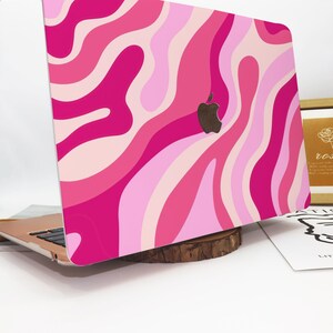 May include: A laptop with a pink and light pink wavy pattern design. The laptop has a black Apple logo. The laptop is resting on a wooden base.