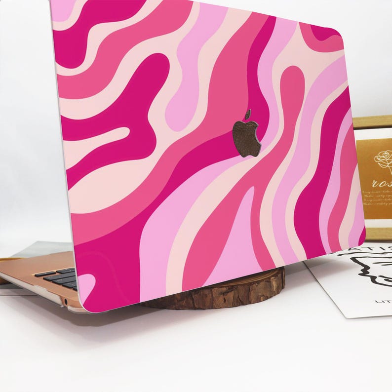 May include: A laptop with a pink and light pink wavy pattern design. The laptop has a black Apple logo. The laptop is resting on a wooden base.
