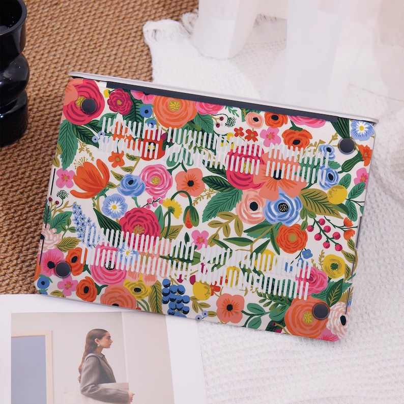 May include: A laptop case with a vibrant floral pattern featuring pink, orange, blue, and yellow flowers on a white background. The case has a protective design with a series of white vertical stripes and black corner accents.