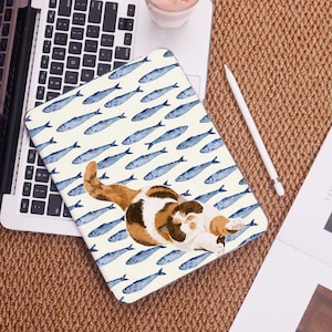 Könnte beinhalten: Eine Laptop-Hülle mit einer Aquarellillustration einer Schildpattkatze auf cremefarbenem Hintergrund mit blauen Fischen. Die Hülle befindet sich auf einem Laptop und einer gewebten Oberfläche.