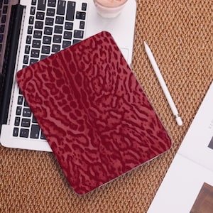 Könnte beinhalten: Eine weinrote Tablethülle mit einem sich wiederholenden Tiermuster liegt auf einem Laptop. Die Hülle befindet sich auf einer braunen, gewebten Oberfläche, mit einem weißen Stift und einem Glas eines rosa Getränks in der Nähe. Der Laptop ist geöffnet.