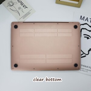 May include: A rose gold laptop bottom case with ventilation slots and four black rubber feet. The text "clear bottom" is at the bottom of the image. The case is on a white surface with a magazine and a framed picture in the background.