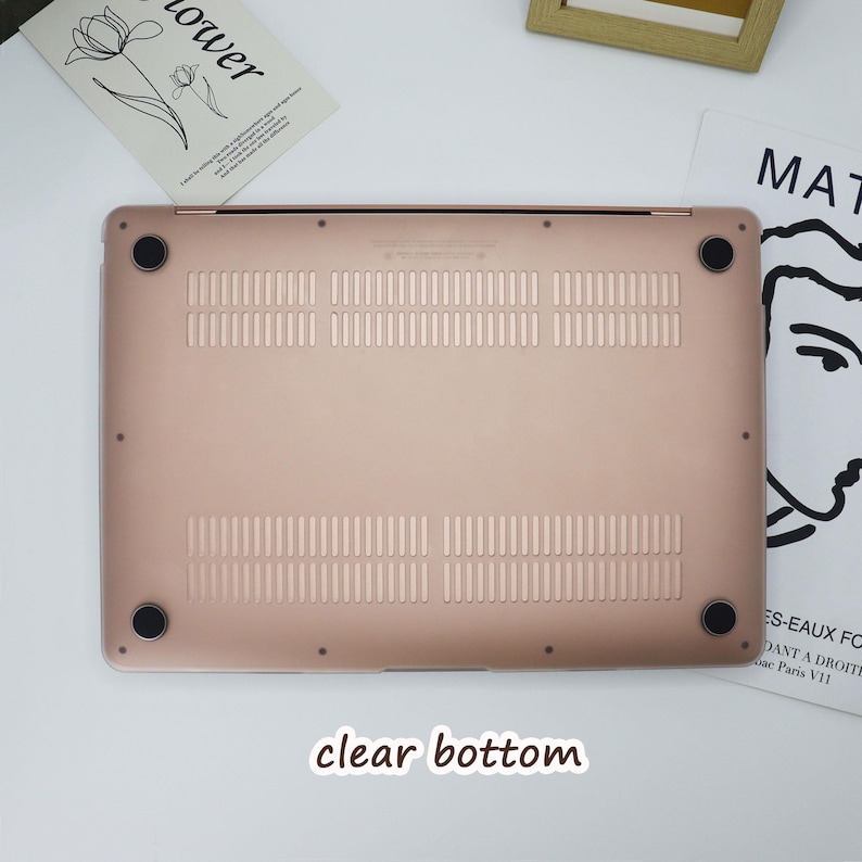May include: A rose gold laptop bottom case with ventilation slots and four black rubber feet. The text "clear bottom" is at the bottom of the image. The case is on a white surface with a magazine and a framed picture in the background.