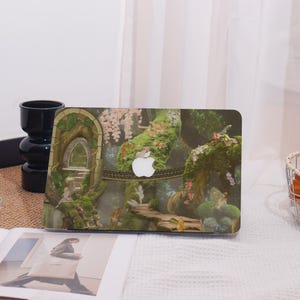 May include: Laptop with a fantasy forest design. The cover shows a bridge, fairies, and lush greenery. The Apple logo is visible. The design includes an archway and a pathway, creating a whimsical scene.