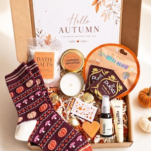 May include: A cardboard gift box containing a pair of maroon socks with pumpkin and mushroom patterns, a bag of bath salts, a pumpkin spiced latte candle, Pukka vanilla chai tea bags, a Cosy vanilla spray, a lip balm, and a heart-shaped soap.