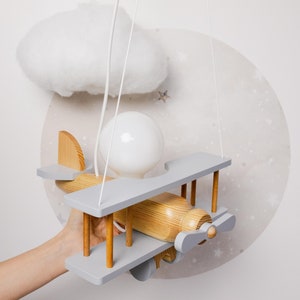 May include: A wooden airplane-shaped ceiling light fixture with a gray and natural wood finish. The light fixture is hanging from a white cord and has a white light bulb. The fixture is suspended in front of a white wall with a white cloud and stars.