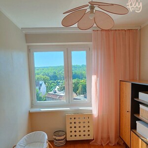 May include: A light-filled nursery with a wicker bassinet, a round jute rug, and a wooden cabinet. The room features a large window with sheer pink curtains and a ceiling fan with petal-shaped blades. The walls are painted a neutral color.