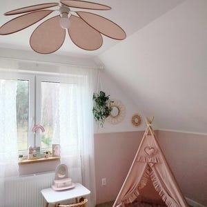 Peut inclure: Chambre d'enfant aux tons roses et blancs. Un ventilateur de plafond en forme de fleur est suspendu au-dessus d'une petite table blanche et d'une chaise en osier. Une tente tipi rose est dans le coin, avec le texte "fly with us".