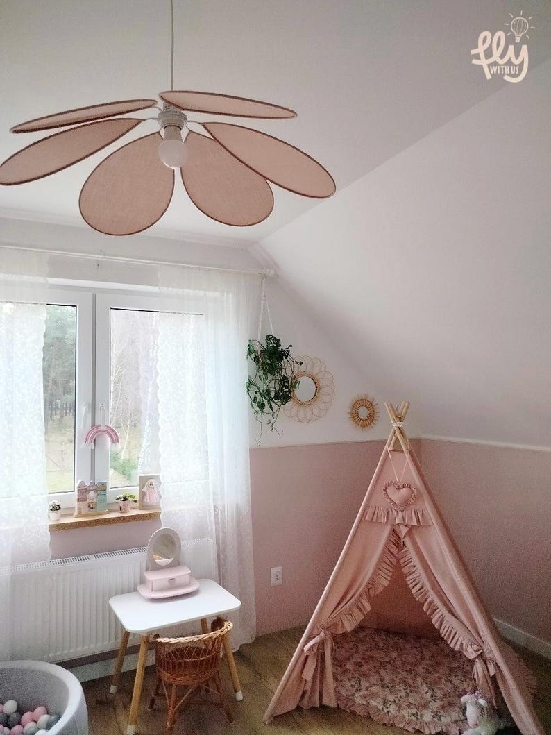 May include: A child's bedroom with a pink and white color scheme. A flower-shaped ceiling fan hangs above a small white table and a wicker chair. A pink teepee tent is in the corner, and the text "fly with us" is in the upper right corner.