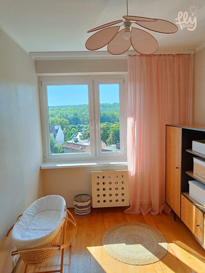 May include: A light-filled nursery with a wicker bassinet, a round jute rug, and a wooden cabinet. The room features a large window with sheer pink curtains and a ceiling fan with petal-shaped blades. The walls are painted a neutral color.