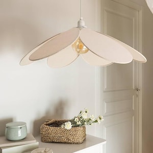 May include: A flower-shaped pendant light with a beige fabric shade and a visible lightbulb. The light hangs above a white table with a woven basket, flowers, and decorative items. The room has a neutral color palette.