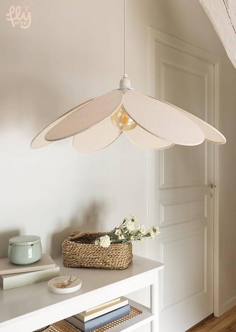 May include: A flower-shaped pendant light with a beige fabric shade and a visible lightbulb. The light hangs above a white table with a woven basket, flowers, and decorative items. The room has a neutral color palette.