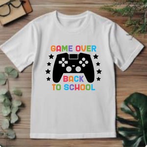May include: White short-sleeved t-shirt with a colorful graphic. The graphic features a black video game controller, the words "GAME OVER" in orange, and "BACK TO SCHOOL" in blue, with stars. The shirt is displayed on a wooden surface.