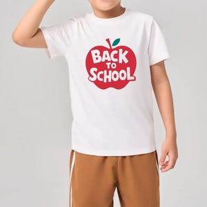 May include: White t-shirt with a red apple graphic and the text "Back to School". The shirt is paired with brown shorts. The outfit is suitable for children.