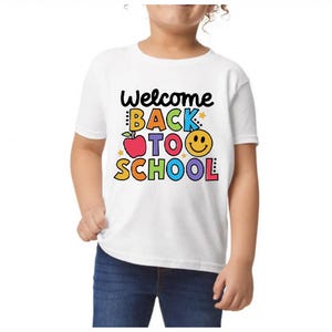 May include: White t-shirt with the text "Welcome Back to School" in colorful letters and a smiling face. The design includes an apple and stars. The shirt is worn by a child.