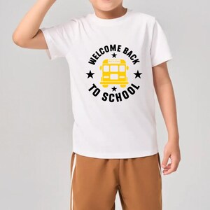 May include: White t-shirt with a yellow school bus graphic and the text "WELCOME BACK TO SCHOOL". The shirt is paired with brown shorts. A back-to-school themed outfit.