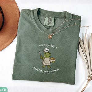 May include: A moss green short-sleeved t-shirt featuring a graphic of a frog wearing a chef hat and apron, holding a whisk and a bowl. The text reads OFF TO HAVE A MENTAL BAKE DOWN. The shirt has a Comfort Colors label indicating size small.