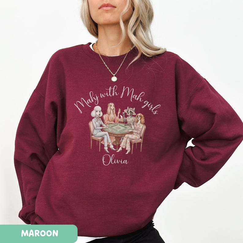 Op de afbeelding: Bordeaux sweatshirt met de tekst "Mahy with Mah girls" en de naam "Olivia". De afbeelding toont vier vrouwen die een spel spelen. Het woord "MAROON" staat onderaan de afbeelding.