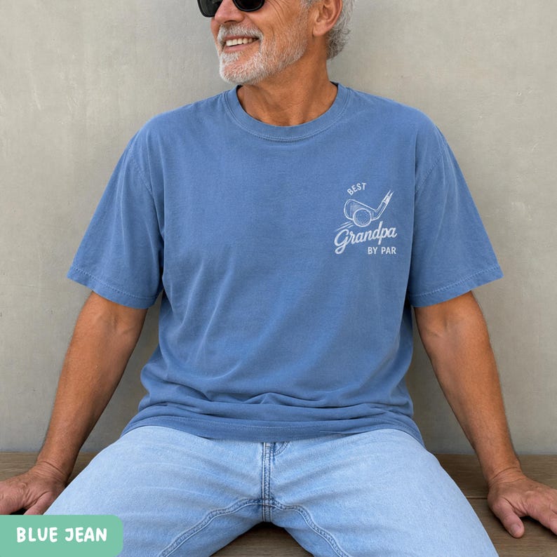 May include: A blue short-sleeved t-shirt featuring a white graphic on the left chest. The graphic depicts a golf club and ball with the text "BEST" above and "Grandpa BY PAR" below. The shirt is paired with light blue denim jeans.