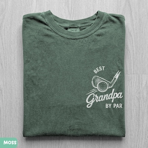 May include: A moss green crew neck t-shirt featuring a white graphic on the lower right side. The graphic depicts a golf club and ball with the text "BEST" above and "Grandpa BY PAR" below. The shirt has a ribbed collar and a visible inner neck label.