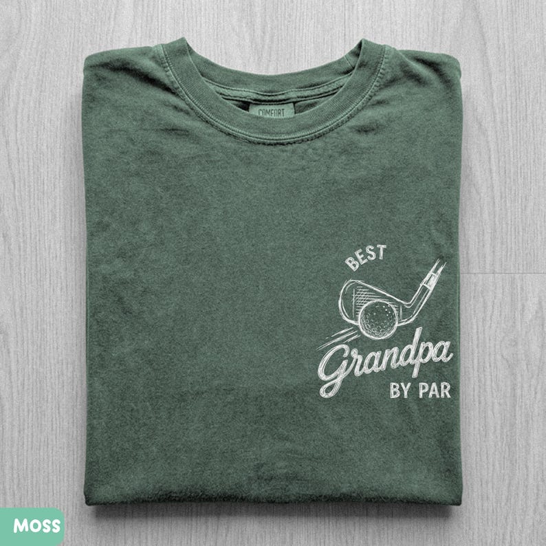 May include: A moss green crew neck t-shirt featuring a white graphic on the lower right side. The graphic depicts a golf club and ball with the text "BEST" above and "Grandpa BY PAR" below. The shirt has a ribbed collar and a visible inner neck label.