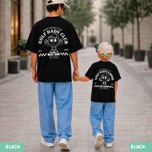 May include: Two black t-shirts with white graphics, one for an adult and one for a child, both with the text "Golf Dads Club" and "Best Dad By Par". The child's shirt says "Golf Dad's Caddie" and "Tiny Golfer". Both are wearing light blue jeans.