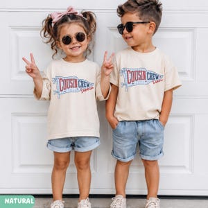 May include: Two children in matching outfits: beige t-shirts and denim shorts. The shirts display a banner design with "COUSIN CREW" and names. One child has a pink bow and sunglasses, the other wears sunglasses. The image is set against a white door.