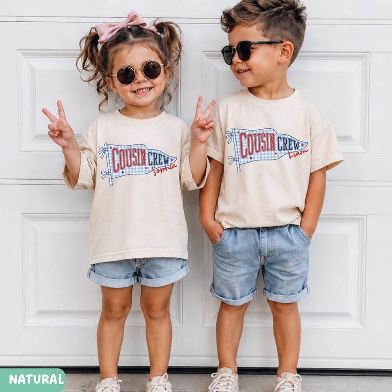 May include: Two children in matching outfits: beige t-shirts and denim shorts. The shirts display a banner design with "COUSIN CREW" and names. One child has a pink bow and sunglasses, the other wears sunglasses. The image is set against a white door.