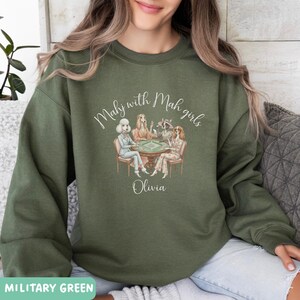 Op de afbeelding: Militairgroene sweatshirt met de tekst "Mahj with Mah girls" en de naam "Olivia". De afbeelding toont vier vrouwen die een spel spelen. De sweatshirt is effen van kleur.