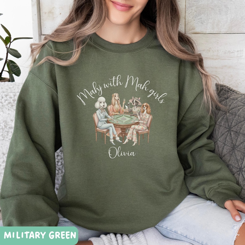 Op de afbeelding: Militairgroene sweatshirt met de tekst "Mahj with Mah girls" en de naam "Olivia". De afbeelding toont vier vrouwen die een spel spelen. De sweatshirt is effen van kleur.