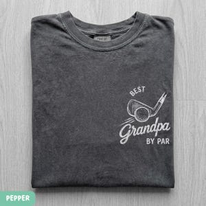 May include: A charcoal gray crew neck t-shirt featuring a white graphic on the chest. The design includes a sketch of a golf club and ball with the text "BEST" above and "Grandpa BY PAR" below the illustration. The shirt has a ribbed collar.