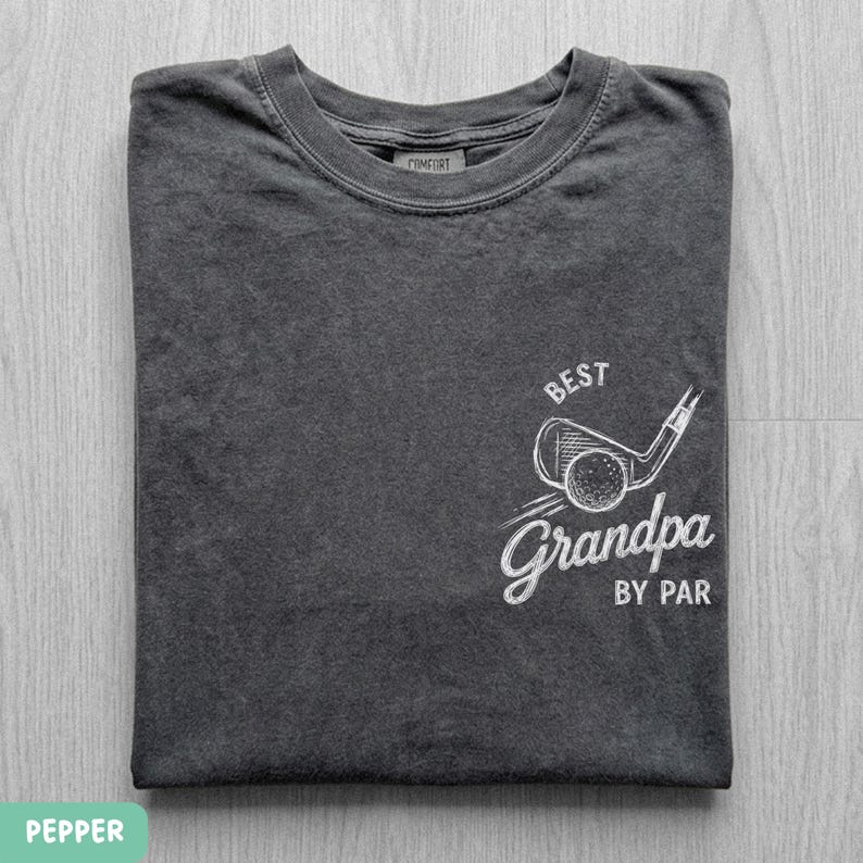 May include: A charcoal gray crew neck t-shirt featuring a white graphic on the chest. The design includes a sketch of a golf club and ball with the text "BEST" above and "Grandpa BY PAR" below the illustration. The shirt has a ribbed collar.
