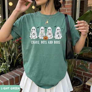 May include: A sage green t-shirt featuring a graphic of four white ghosts holding mahjong tiles, a small orange jack-o'-lantern, and the text "CRAKS, DOTS AND BOOS" printed in white. The shirt has a crew neckline and short sleeves.