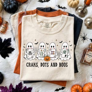 May include: A cream-colored t-shirt featuring a graphic of four white ghosts holding mahjong tiles, a small orange pumpkin, spiderwebs, and bats. The text "CRAKS, DOTS AND BOOS" is printed in black below the ghosts. The shirt has a crew neckline.