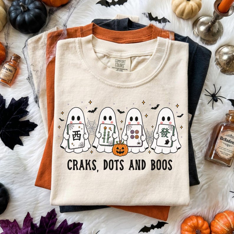 May include: A cream-colored t-shirt featuring a graphic of four white ghosts holding mahjong tiles, a small orange pumpkin, spiderwebs, and bats. The text "CRAKS, DOTS AND BOOS" is printed in black below the ghosts. The shirt has a crew neckline.