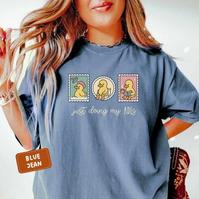 May include: Blue jean colored t-shirt with a graphic of three postage stamp-like images featuring a yellow duck. The duck is shown showering, on the toilet, and reading. The text "just doing my ADLs" is below the images.