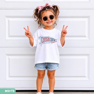 May include: A child wearing a white t-shirt with the text "Cousin Crew Sophia" printed on it, paired with denim shorts. The child is wearing sunglasses and a pink bow. The child is giving a peace sign.