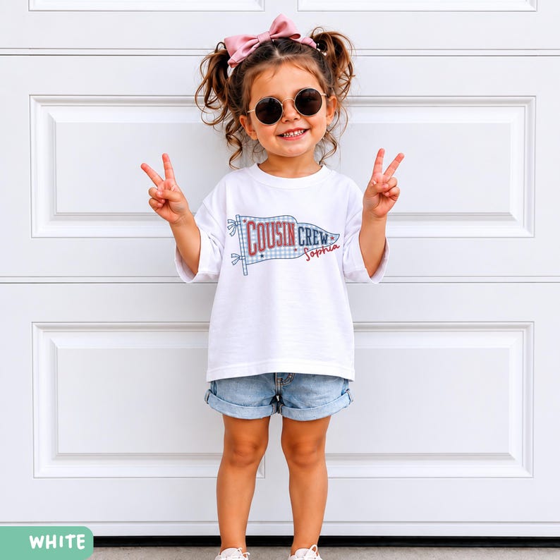 May include: A child wearing a white t-shirt with the text "Cousin Crew Sophia" printed on it, paired with denim shorts. The child is wearing sunglasses and a pink bow. The child is giving a peace sign.