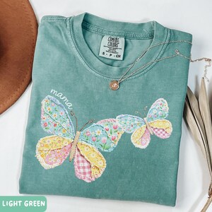 May include: Light green t-shirt with a butterfly design. The butterfly wings are made of floral and gingham patterns. The word "mama" is written above the butterfly. A silver necklace with a pendant is also visible.